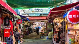 Ein belebter Markt mit bunten Ständen, die eine Vielzahl von Waren und Souvenirs anbieten, unter einem grünen Sonnenschutz.