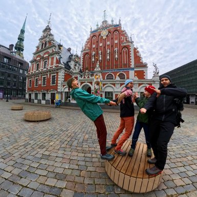 Vier Personen stehen auf einer Holzplattform und posieren fröhlich vor einem historischen Gebäude in Riga.