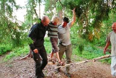 Ein Junge balanciert auf einem Seil, während zwei Erwachsene ihn unterstützen und lachen, umgeben von Bäumen und Natur.