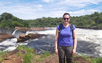 Eine Frau steht lächelnd am Ufer des Murchison Falls, umgeben von üppiger Natur und sprudelndem Wasser.