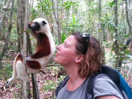 Eine Frau steht in einem dichten Wald und schaut liebevoll zu einem Sifaka, der an einem Baumstamm klettert.