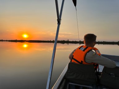 Ein Junge in einer orangefarbenen Schwimmweste sitzt in einem Boot und beobachtet den Sonnenuntergang über einem ruhigen Gewässer.