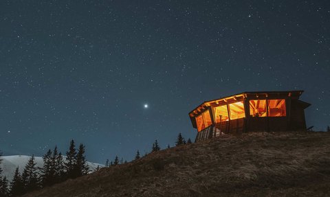 Eine beleuchtete Hütte steht auf einem Hügel, umgeben von dunklem Himmel und funkelnden Sternen.