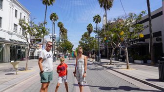 Eine Familie steht fröhlich auf der Santa Monica Promenade, umgeben von Palmen und Geschäften unter blauem Himmel.