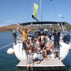 Eine fröhliche Gruppe von Menschen winkt von einem Segelboot aus, umgeben von klarem Wasser und sanften Hügeln.