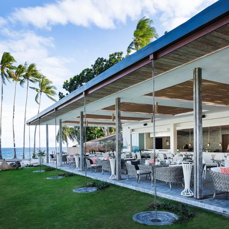 Ein modernes Restaurant am Strand mit stilvollen Möbeln und Blick auf das Meer, umgeben von hohen Palmen und klarem Himmel.