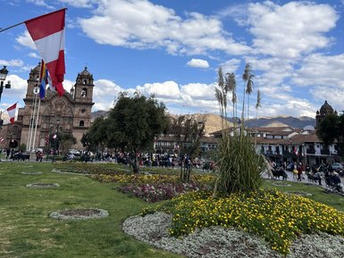 Im Plaza de Armas in Cusco wehen die peruanischen Flaggen im Wind, umgeben von bunten Blumenbeeten und einem strahlend blauen Himmel.