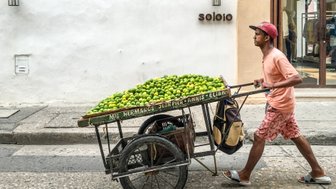 Ein Mann schiebt einen Wagen voller frischer Limetten durch eine belebte Straße in Cartagena, umgeben von weißen Wänden.