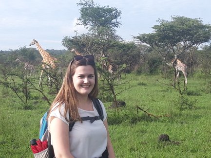 Eine junge Frau lächelt vor einer grünen Landschaft mit mehreren Giraffen im Hintergrund, die an Bäumen fressen.