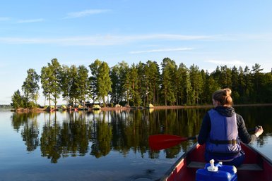 Eine Person paddelt in einem Kanu auf einem ruhigen See, umgeben von grünen Bäumen und reflektierenden Wasserflächen.