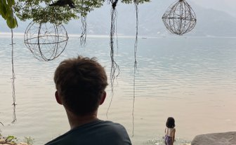 Ein Junge sitzt am Ufer des Phewa-Sees und beobachtet ein Mädchen, das am Wasser spielt, umgeben von grünen Bäumen.