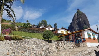 Bunte Häuser mit blühenden Pflanzen vor dem beeindruckenden Felsen El Peñol unter einem strahlend blauen Himmel.