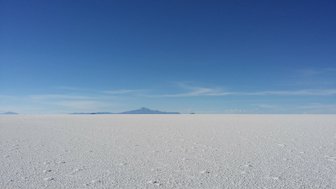 Eine weite, weiße Salzfläche erstreckt sich bis zum Horizont, unter einem strahlend blauen Himmel mit wenigen Wolken.