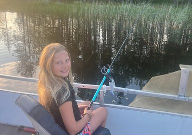 Ein Mädchen sitzt auf einem Boot und angelt, umgeben von Bäumen und ruhigem Wasser, mit einem Lächeln auf dem Gesicht.
