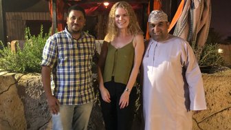 Drei Personen stehen lächelnd vor einem festlich dekorierten Zelt mit orangefarbenen Stoffen und sanfter Beleuchtung.
