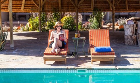 Eine Frau in Sonnenbrille und Hut entspannt am Pool, während sie ein Buch liest und ein Getränk auf einem Tisch hat.