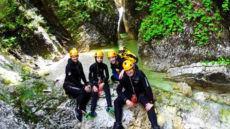 Fünf Personen in Neoprenanzügen und Helmen posieren fröhlich an einem Wasserfall in einer grünen Schlucht.