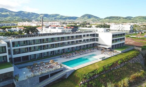 Ein modernes Hotel mit einem großen Pool und Liegen, umgeben von grünen Hügeln und einer malerischen Landschaft.