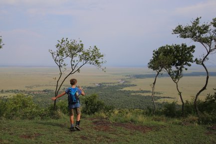 Ein Junge steht auf einem Hügel und blickt über die weite, grüne Landschaft der Masai Mara, umgeben von Bäumen.
