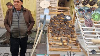 Ein Mann steht an einem Marktstand, umgeben von handgefertigten Holzgegenständen und bunten Schalen im Hintergrund.