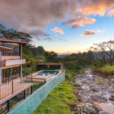 Ein modernes Haus mit großen Fenstern bietet einen Blick auf einen ruhigen Fluss und eine malerische Landschaft bei Sonnenuntergang.
