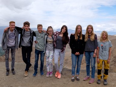 Eine Gruppe von acht Jugendlichen posiert fröhlich auf einem Berggipfel mit einer beeindruckenden Aussicht im Hintergrund.