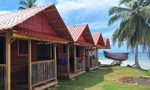 Verschiedene Holzbungalows mit roten Dächern stehen auf einer grünen Wiese am Strand, umgeben von Palmen und blauem Wasser.
