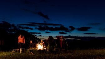 Eine Gruppe von Menschen versammelt sich um ein Lagerfeuer, während der Himmel in der Dämmerung leuchtet.