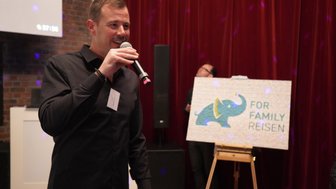 Ein Mann in einem schwarzen Hemd spricht mit einem Mikrofon, während er auf eine bunte Elefantenmalerei zeigt.