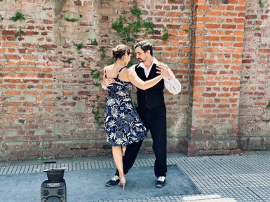 Ein Paar tanzt leidenschaftlich Tango vor einer roten Ziegelmauer, umgeben von einer lebhaften Atmosphäre.