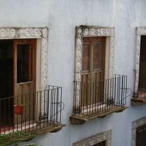 Drei Fenster mit kunstvollen Rahmen und schmiedeeisernen Balkonen, umgeben von grünen Pflanzen und einer hellen Wand.