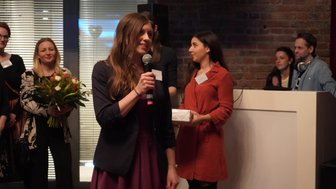 Eine Frau spricht mit einem Mikrofon, während sie in einem festlichen Raum steht, umgeben von anderen Anwesenden.