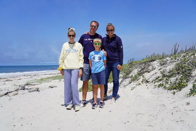 Eine Familie steht am Strand, umgeben von Sand und Meer, und lächelt fröhlich in die Kamera.