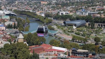 Eine Seilbahn schwebt über die Stadt Tiflis, mit Blick auf den Fluss und moderne Architektur im Hintergrund.