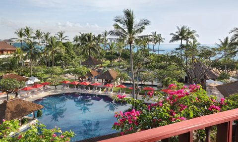 Ein Blick auf den Pool des Nusa Dua Beach Hotels, umgeben von Palmen und bunten Blumen, mit Sonnenliegen und roten Sonnenschirmen.