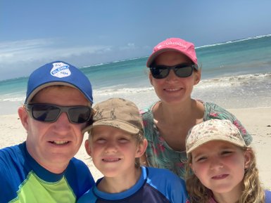Eine Familie posiert am Strand, umgeben von türkisfarbenem Wasser und einem klaren Himmel, alle lächeln glücklich.