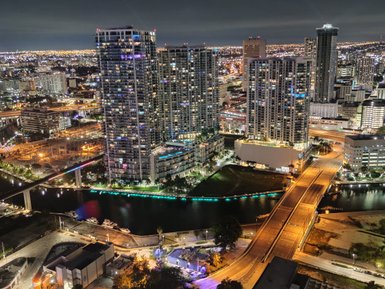 Die nächtliche Skyline von Miami erstrahlt in bunten Lichtern, während der Fluss ruhig darunter fließt.