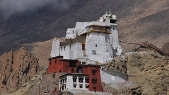 Ein beeindruckendes Kloster thront auf einem steilen Felsen, umgeben von majestätischen Bergen und einem klaren Himmel.