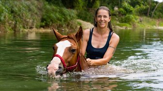 Eine Frau reitet auf einem schwimmenden Pferd in einem klaren, grünen Gewässer und lächelt glücklich in die Kamera.
