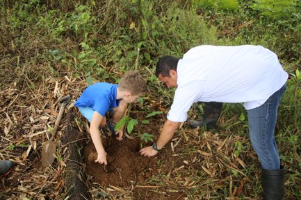Ein Junge und ein Erwachsener pflanzen gemeinsam einen kleinen Baum in der Erde, umgeben von üppigem Grün und Pflanzenresten.