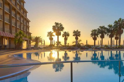Ein atemberaubender Sonnenuntergang spiegelt sich im glitzernden Pool eines Hotels, umgeben von Palmen und Liegen.
