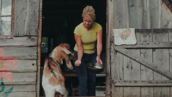 Eine Frau steht an der Tür einer Holzhütte und füttert eine Ziege mit einer Schüssel in der Hand.