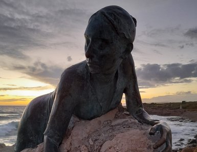 Eine bronzene Skulptur einer Frau, die auf einem großen Stein sitzt, während die Wellen im Hintergrund sanft brechen.