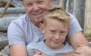 Ein lächelnder Mann umarmt einen Jungen, während sie auf einem Kiesstrand sitzen, umgeben von Holzstämmen.