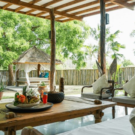 Ein einladendes Wohnzimmer im Freien mit Holzdekor, umgeben von üppigem Grün und einem traditionellen Pavillon im Hintergrund.