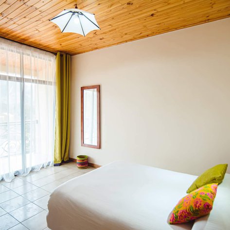 Ein helles Hotelzimmer mit einem großen Fenster, das Tageslicht hereinlässt, und einem gemütlichen Bett mit bunten Kissen.