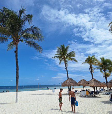 Zwei Jungen gehen barfuß über den weißen Sandstrand, umgeben von Palmen und Sonnenschirmen, während das Meer im Hintergrund glitzert.