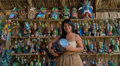 Eine Frau in traditioneller Kleidung hält ein Musikinstrument und lächelt vor einer Wand voller bunter handgefertigter Masken.