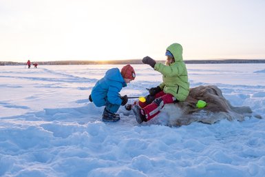 Zwei Kinder spielen auf dem gefrorenen See, während die Sonne hinter den Bäumen untergeht und die Landschaft in warmes Licht taucht.