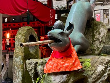 Eine bronzeartige Statue eines Hundes mit einem Stock im Mund, gekleidet in ein leuchtend rotes Tuch, umgeben von Steinen.